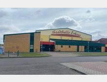 Außenansicht der Stadthalle Erkner, einem modernen Veranstaltungsgebäude mit gelber und brauner Klinkerfassade, einem grün abgesetzten Flachdachbereich und roten Akzenten über dem Eingang. Im Vordergrund sind eine Straße, Gehwege und ein Fahrradständer zu sehen.