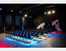 Veranstaltungsraum mit mehreren Reihen schwarzer Stühle, die auf eine Bühne mit einer weißen, geschwungenen Wand ausgerichtet sind. Der Raum ist dunkel gehalten und durch bunte Lichteffekte in Blau und Rot beleuchtet; im Hintergrund befinden sich Glasbausteine und technische Elemente.