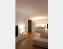 Minimalistisch eingerichtetes Wohnzimmer mit hellen Wänden und Holzboden, sichtbar sind ein beiger Sessel, ein TV auf einem niedrigen weißen Sideboard mit einer Lampe, sowie eine offene, moderne Küche im Hintergrund mit weißen Hochglanzschränken.