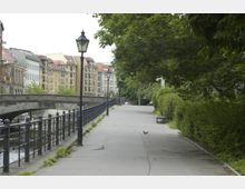 Uferweg entlang eines Flusses mit schmiedeeisernem Geländer, Straßenlaternen und einer steinernen Brücke im Hintergrund. Auf der linken Seite stehen Gebäude mit roten Ziegeldächern, während die rechte Seite von Bäumen und Sträuchern gesäumt ist.