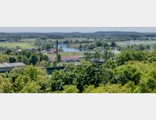 Panoramaansicht einer ländlichen Gegend mit dichtem Baumbestand im Vordergrund, vereinzelten Gebäuden, einer Fabrik mit einem hohen Schornstein in der Mitte und einem Fluss, der sich durch die Grünflächen schlängelt. Im Hintergrund sind Wiesen, Wälder und sanfte Hügel unter einem klaren Himmel zu sehen.