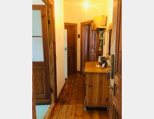 Schmaler Hausflur mit Holzdielenboden und holzverkleideten Türen. Auf der rechten Seite steht eine hölzerne Kommode mit Deko, darüber ein Wandregal mit Körben und Taschen.