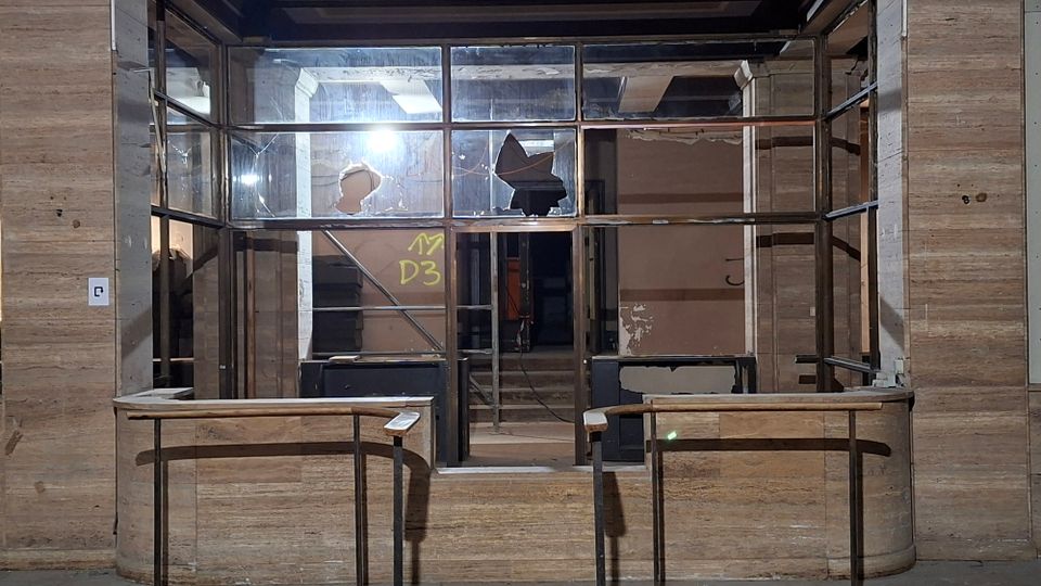 Verlassener Empfangs- oder Kassenbereich in einem älteren Gebäude mit Holzverkleidung, zwei gebogenen Tresen und einer großen Glaswand mit Metallrahmen und teilweise zerbrochenen Scheiben. Dahinter sind ein Treppenaufgang, freiliegende Konstruktionselemente und abgenutzte Wände in einem dunklen Innenraum zu sehen.