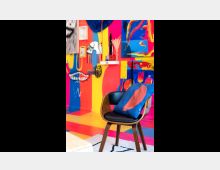 Ein bunter Raum mit einer Wand aus abstrakten Gemälden in kräftigen Farben wie Rot, Blau und Gelb. Im Vordergrund steht ein Stuhl mit schwarzem Sitz und einem blauen Kissen mit einem roten Motiv, der auf einem hellen Teppich positioniert ist.