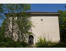 Ein schlichter, rechteckiger Betonbau mit hellem Putz, kleinen rechteckigen Fenstern und einem Rundbogen-Eingang. Der Bau ist von Bäumen und dichter Vegetation umgeben, während der blaue Himmel im Hintergrund zu sehen ist.