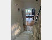 Blick von einer weißen Treppe mit dunklem Handlauf in einen Flur mit gemustertem Fliesenboden. Im Hintergrund ist ein Essbereich mit Holzboden, einem Tisch, Stühlen und bodentiefen Fenstern zu sehen.