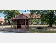 Kleine Bushaltestelle aus Fachwerk mit Ziegeldach und Sitzbank, umgeben von Bäumen. Im Hintergrund stehen ein großes, mit Efeu bewachsenes Gebäude mit weißen Sprossenfenstern und blauer Eingangstür sowie eine gepflasterte Straße.