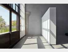 Minimalistischer Innenraum mit einer grauen Betonwand, einem großen Fenster mit schwarzen Rahmen und weißen Vorhängen sowie einer weißen Trennwand, auf der das Sonnenlicht ein rechteckiges Muster erzeugt. Durch das Fenster ist ein gepflegter Außenbereich mit Rasen und Bäumen zu sehen.