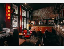 Innenraum einer Bar mit hohen Fensterfronten, sichtbarer Backsteinwand und dunkler Decke mit Spotbeleuchtung. Entlang der Bar und einer gepolsterten Sitzbank stehen kleine Tische mit gemischten Stühlen; Spiegel und Hängelampen rahmen den Tresenbereich.