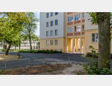 Wohnblock mit heller Fassade in Gelb- und Weißtönen, umgeben von einem begrünten Gartenbereich mit Bäumen und Sträuchern. Der Gebäudeteil zeigt einen Durchgang mit Säulen, im Hintergrund sind weitere Wohngebäude sichtbar.