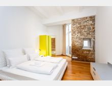 Helles Schlafzimmer mit Doppelbett, weißer Bettwäsche und Holzboden, kombiniert mit einer freiliegenden Backsteinwand mit kleinem Fenster. Ein auffälliger gelber Kleiderschrank steht neben bodentiefen Fenstern mit Vorhängen, die viel Tageslicht in den Raum lassen.