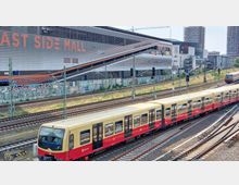 Ein gelb-roter Berliner S-Bahn-Zug fährt auf Schienen vor der "East Side Mall", einem modernen Einkaufszentrum mit auffälliger Fassade. Im Hintergrund sind graffitibedeckte Mauern, mehrere Bahngleise und hohe Gebäude sichtbar.