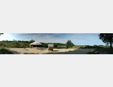 Panoramaaufnahme eines Sandstrandes an einem See, umgeben von Bäumen und dichter Vegetation. Im Vordergrund stehen ein Strandkorb, ein Sonnenschirm und Liegestühle, mit klarem Himmel im Hintergrund.