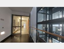 Ein heller, moderner Gebäudeflur mit gläsernem Aufzug auf der rechten Seite, Metallgeländer mit Holzhandlauf und einer Reihe von Türen und Stühlen entlang der linken Wand. Der Flur führt in einen weiteren gut beleuchteten Bereich mit Glasfenstern.