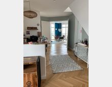 Wohnzimmeransicht in einer modernen Wohnung mit hellem Fischgrätenparkett. Im Vordergrund ist eine offene Küche mit Theke zu sehen, im Hintergrund ein Essbereich mit blau gestrichener Wand, großen Fenstern und einer Hängelampe.