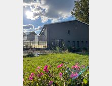 Ein modernes Gebäude mit dunkler Fassade steht neben einem Gewächshaus. Im Vordergrund befinden sich eine grüne Rasenfläche mit blühenden pinken Blumen sowie Gartenmöbel unter sonnigem Himmel mit Wolken.