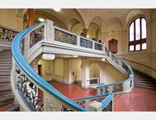 Treppenhaus in einem historischen Gebäude mit geschwungenen Treppen und kunstvoll verzierten Schmiedeeisengeländern. Die Wände sind mit detaillierten Stuckornamenten versehen, und große Rundbogenfenster lassen Tageslicht in das Innere.