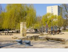Spielplatz mit Sandboden, Holzklettergeräten und Betonstrukturen, umgeben von hängenden Weidenbäumen; im Hintergrund ein mehrstöckiges Wohngebäude.