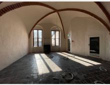 Raum mit gewölbter Decke und Ziegelbögen, rohen weißen Wänden und einem Boden aus dunklen Steinfliesen. Zwei große Fenster lassen Tageslicht herein, und auf dem Boden liegen lose Kabel.