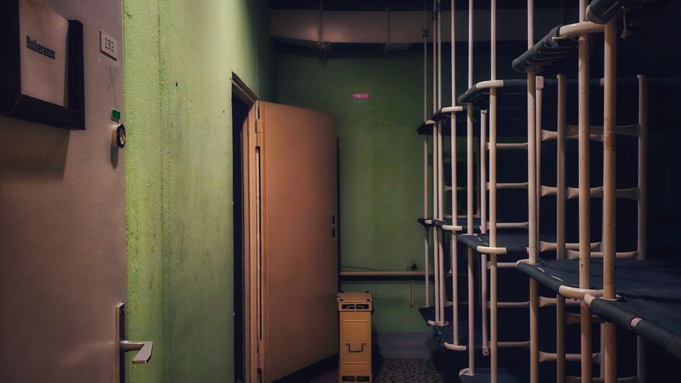 Schmaler Innenflur mit grün gestrichenen Wänden, Neon-Deckenleuchte und gemustertem Bodenbelag. Rechts stehen hohe, metallene Rohrrahmen-Regale, links befinden sich Türen und Schaltschränke; am Ende steht ein kleiner Wagen.