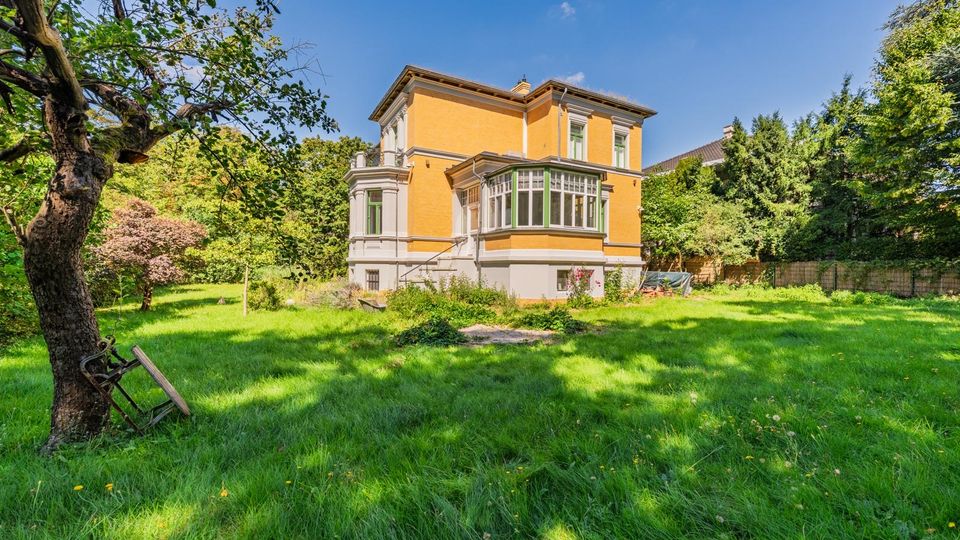 Freistehende, zweigeschossige Villa mit gelb-orangefarbener Fassade, grauem Sockel und verglastem Erker, umgeben von einem großen, grünen Garten. Im Vordergrund liegt eine Rasenfläche mit altem Baum; im Hintergrund stehen dichtes Buschwerk und hohe Bäume unter blauem Himmel.