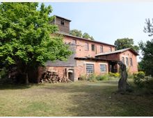 Ziegelsteingebäude mit Holzanbau, mehrere Fenster und ein Wellblechdach, umgeben von Bäumen und Wiese. Im Vordergrund ein großer Baum, ein Holzstapel und eine Statue in einer Gartenlandschaft.