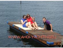 Drei Personen befinden sich auf einem kleinen, rechteckigen Holzsteg auf einem Gewässer. Zwei Personen sitzen auf einer leicht gepolsterten Unterlage in der Mitte des Stegs, während eine dritte am Rand im Wasser ist.