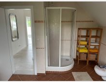 Ein Badezimmer mit einer Eckdusche, hellen Fliesen mit roten Akzenten und einem braunen Fliesenboden. Rechts steht ein Holzregal mit gefalteten Handtüchern und Körben, daneben eine Toilette; links führt eine Tür in einen angrenzenden, hellen Raum mit einem Spiegel.