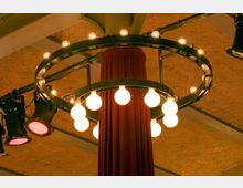 Eine rote, mit Stoff verkleidete Säule wird von einem kreisförmigen Kronleuchter mit zahlreichen Glühbirnen umgeben, der an einer metallischen Aufhängung befestigt ist. Der Hintergrund zeigt eine ziegelartige Decke mit sichtbaren Trägern und zusätzlich montierten Bühnenscheinwerfern.