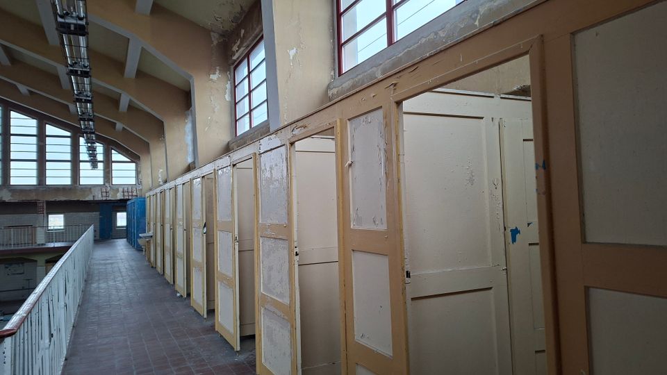 Ein verlassener Gebäudeinnenraum mit verwitterten Umkleidekabinen aus Holz, abblätternder Farbe und hohen Fenstern mit roten Rahmen, die Tageslicht hereinlassen. Die Decke hat freiliegende Träger, und rechts verläuft ein Gang entlang einer offenen Galerie mit Geländer.