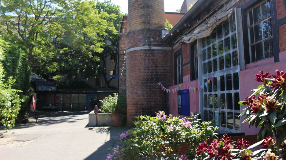 Innenhof eines Backstein-Industriegebäudes mit hohem zylindrischem Schornstein und großen, gerasterten Fensterfronten an einer rosafarbenen Fassade. Im Vordergrund stehen blühende Sträucher und Pflanzenkübel, daneben führt eine asphaltierte Zufahrt unter hohen Bäumen entlang.