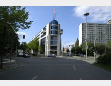 Straßenszene mit moderner Büroarchitektur, einem markanten Gebäude mit weißen Fassaden und großen Glasflächen in der Bildmitte. Umgeben von Bäumen, weiteren Wohn- und Bürogebäuden sowie einer Kreuzung mit Verkehrsampeln.