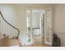 Helles Wohnzimmer mit einer weißen Wendeltreppe und einer geöffneten Glastür, die zu einer modernen Küche mit Holzfußboden führt; im Vordergrund ein geflochtener Sessel und eine grünliche Steinskulptur.