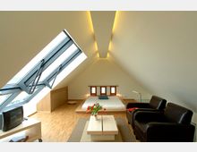 Ein modernes, minimalistisch eingerichtetes Schlafzimmer in einem Dachgeschoss mit schrägen Wänden und großen Dachfenstern. Der Raum verfügt über ein niedriges Bett, einen Tisch mit Blumen, zwei Ledersessel und einen Holzboden.