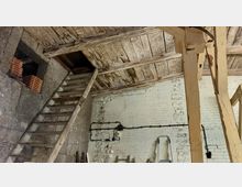Innenansicht eines rustikalen Scheunen- oder Dachbodens. Sichtbare Holzbalken in der Decke, eine altersschwache hölzerne Treppe führt zu einer darüberliegenden Öffnung, und die Wände bestehen aus verputzten Ziegeln mit sichtbaren Kabelinstallationen.