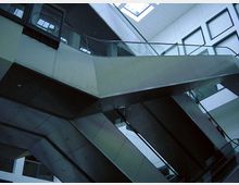 Eine moderne Innenansicht mit mehreren Rolltreppen, die in verschiedene Etagen führen. Das Gebäude hat ein geometrisches Design mit Glasgeländern, Metalloberflächen und einem Oberlicht, das natürliches Licht hereinlässt.