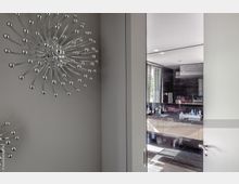 Ansicht eines offenen Badezimmers mit dunklen, glänzenden Wand- und Bodenfliesen, einem großen Spiegel und einem Waschtisch mit Pflegeprodukten. Im Vordergrund eine Wand mit silbernen, sternförmigen 3D-Dekorationen.