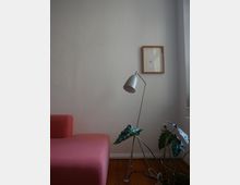 Minimalistisch eingerichteter Wohnraum mit einem roten Sofa, einer silbernen Stehlampe, einer Zimmerpflanze auf Holzboden und einem kleinen gerahmten Bild an einer weißen Wand.