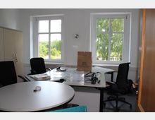 Ein kleines Büro mit zwei großen Fenstern, die auf grüne Bäume blicken. Im Raum befinden sich ein Schreibtisch mit Büromaterialien, ein runder Besprechungstisch, mehrere Bürostühle und ein Holzschrank an der linken Wand.