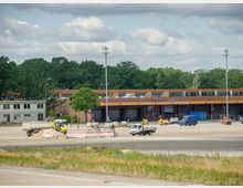 Großes freies Gelände mit Baustellenfahrzeugen, Absperrungen und Sandhaufen, im Hintergrund ein flaches Gebäude mit brauner Fassade und großen Fenstern. Der Bereich ist von Bäumen umgeben, und im Vordergrund sind Grünflächen zu erkennen.