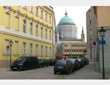 Straßenszene in einer Altstadt mit gepflastertem Straßenbelag, flankiert von einem gelben historischen Gebäude links und modernen Gebäuden rechts. Im Hintergrund ist eine große Kuppel mit Gerüsten sichtbar, vermutlich eine Kirche oder ein öffentliches Gebäude.
