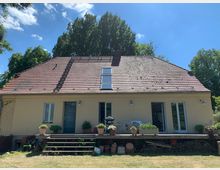 Ein schlichtes Einfamilienhaus mit gelber Fassade und einem steilen Ziegeldach, das eine Dachfensteröffnung aufweist. Vor dem Haus befindet sich eine Holzterrasse mit Pflanzenkübeln, Gartenmöbeln und gestapeltem Brennholz, umgeben von Rasen und hohen Bäumen im Hintergrund.