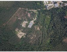 Luftaufnahme eines Waldgebiets mit einer großen Lichtung, mehreren rechteckigen Gebäuden und unbefestigten Wegen. Am oberen Bildrand ist ein angrenzender Industrie- oder Gewerbekomplex mit Hallen und Zufahrten zu sehen.