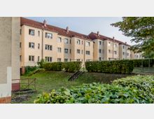 Mehrfamilienhäuser mit gelb-beigen Fassaden und roten Ziegeldächern, umgeben von gepflegten Büschen, Grünflächen und Bäumen; im Vordergrund führt eine kleine Treppe durch die Hecke.