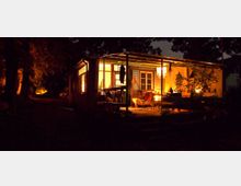 Holzhütte mit überdachter Veranda, beleuchtet durch warmes Licht. Die Umgebung ist dunkel, mit Bäumen und leicht erkennbaren Schatten im Hintergrund.