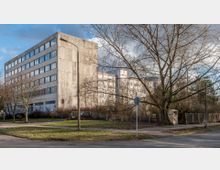 Betongebäude mit mehreren Etagen und rechteckigen Fenstern, umgeben von Bäumen ohne Laub und einer Grünfläche im Vordergrund. Im Vordergrund befinden sich auch eine Straße, Gehwege, Straßenschilder und ein Strommast.