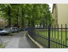 Ein Gehweg mit Kopfsteinpflaster und parkenden Autos entlang einer von Bäumen gesäumten Straße. Rechts im Bild ein gelbes Gebäude und ein schwarzer Metallzaun, der eine Grünfläche umgibt.