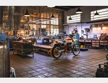 Innenraum einer Werkstatt mit einer Enduro-Motorrad in der Mitte. Der Raum ist mit Metallwerkbänken, Werkzeugen an Wänden, Hängelampen und großen Fenstern ausgestattet, die für eine industrielle Atmosphäre sorgen.