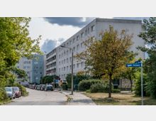 Wohngebiet mit mehrstöckigen, hellen Apartmentgebäuden, an einer schmalen Straße mit geparkten Autos. Bäume und Grünflächen säumen den Gehweg, und ein Straßenschild mit der Aufschrift „An der Schleuse“ ist erkennbar.