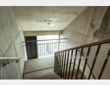 Betontreppe in einem modernen Treppenhaus mit schlichten Wänden aus Sichtbeton, schwarzen Metallgeländern und Holzhandläufen. Ein großes Fenster im Hintergrund lässt Tageslicht einfallen.